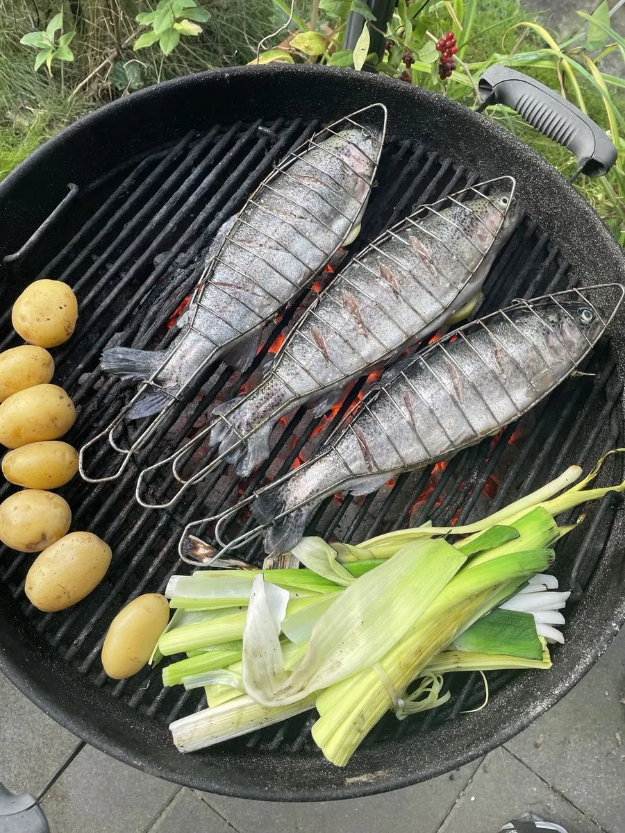 Drei ganze Fische in Grillzangen auf dem Rost, daneben Kartoffeln und aufgeschnittener Lauch