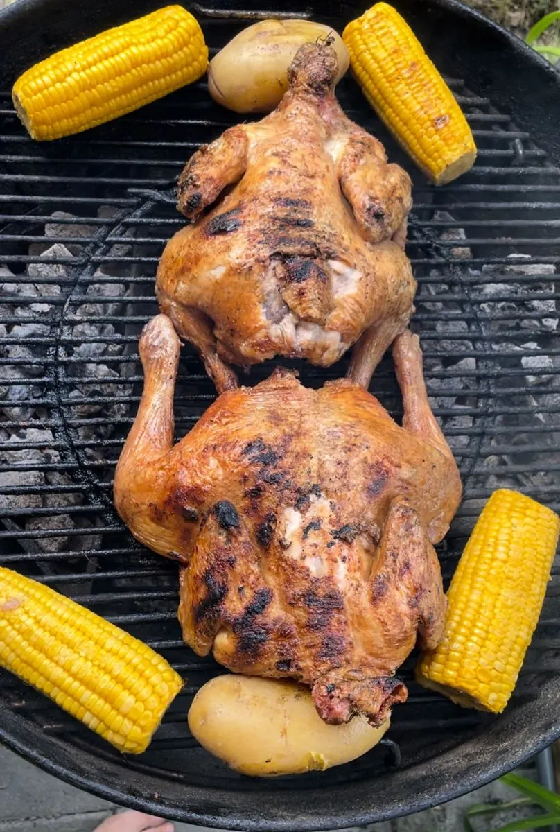 Zwei ganze Hähnchen auf dem Rost, ringsum Maiskolben und Kartoffeln